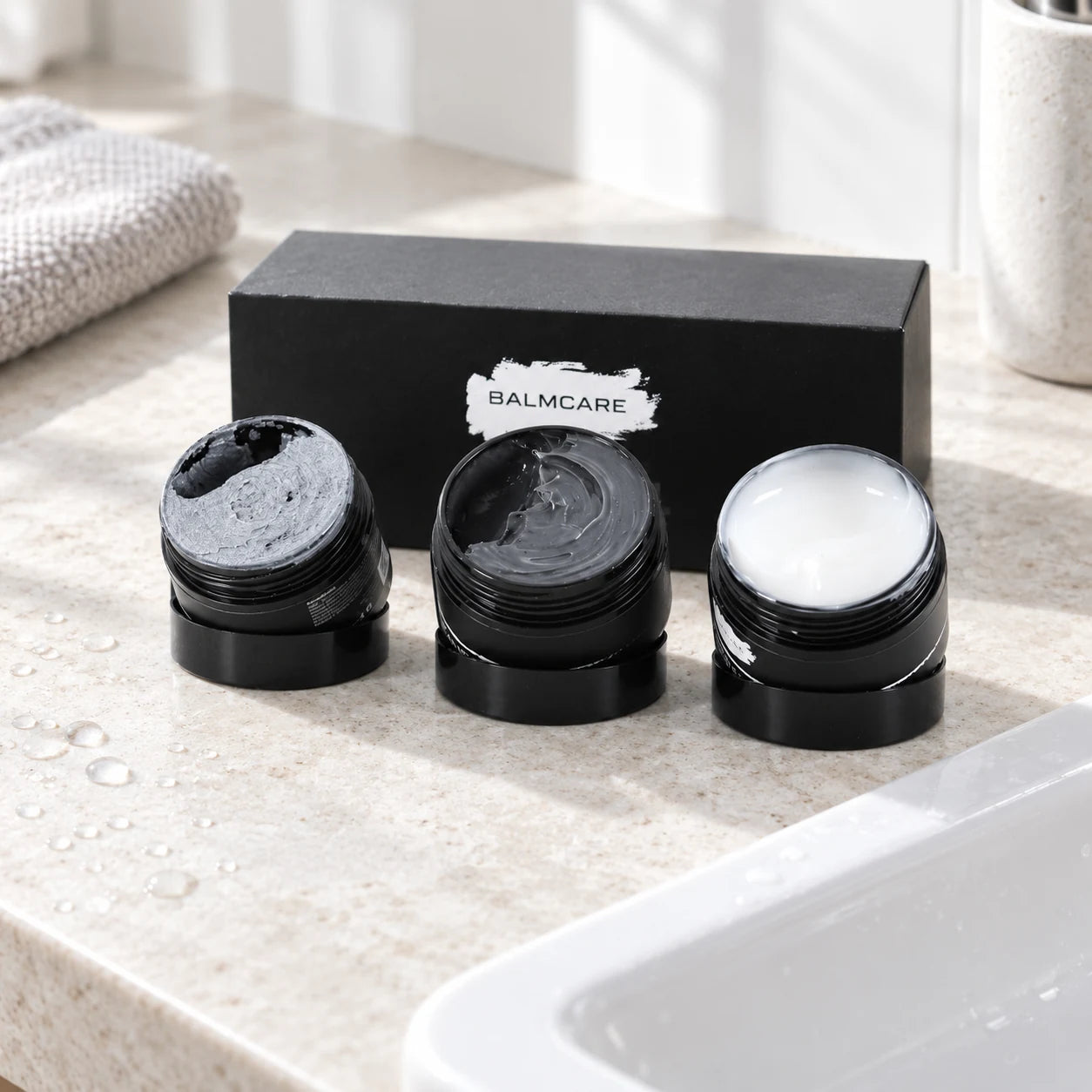 Drie geopende BalmCare huidproducten met grijze scrub, charcoal cleanser en witte moisturizer bij een lichte stenen wastafel